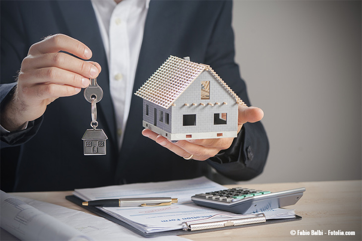 Ein Mann sitzt im Anzug am Schreibtisch. Vor ihm Unterlagen und Taschenrechner, in der einen Hand hält er einen Schlüssel mit einem Schlüsselanhänger in Haus-Form und in der anderen ein Modell-Haus.