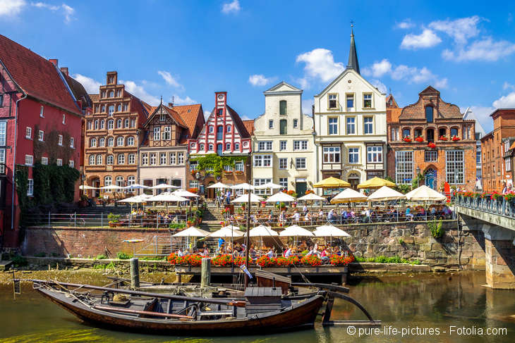 Blick auf die historischen Häuserfassaden vom Hafen aus. Davor ein altes Boot im Wasser und in der Mitte viele Sonnenschirme kleiner Cafés.