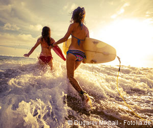 Zwei junge Frauen laufen mit einem Surfbrett unter dem Arm ins Meer