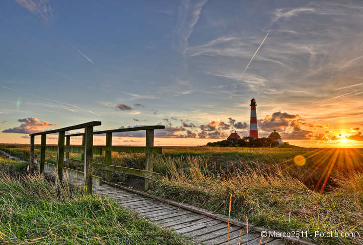 Eine schmale Holzbrücke in einer Gräserlandschaft. Im Hintergrund ein Leuchtturm und der Sonnenuntergang.