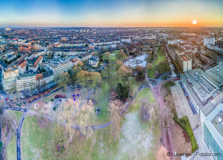 There are trees and a park in the foreground and buildings and streets in the background.